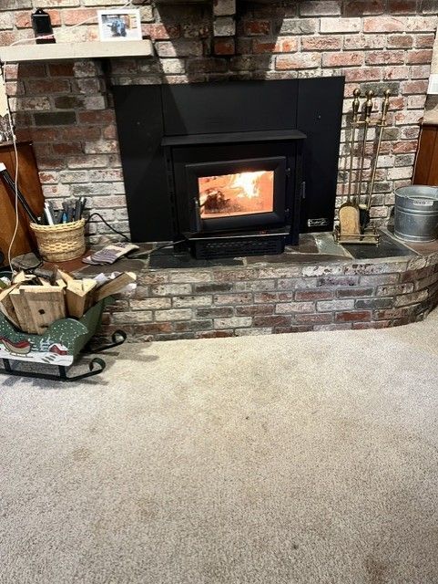 Brick fireplace with a lit wood stove insert on a raised hearth in a carpeted living room