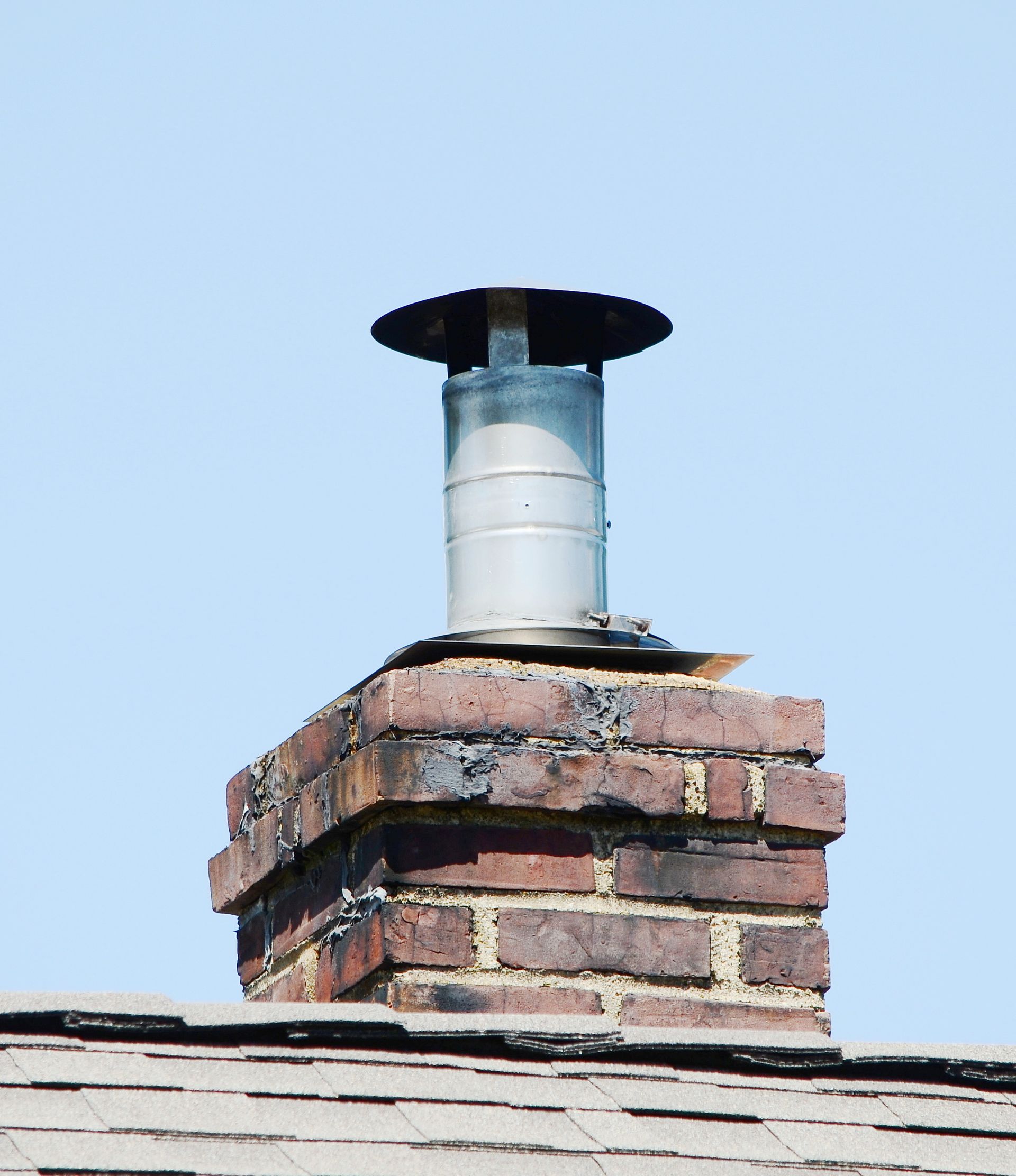 Brick chimney with a silver vent pipe and black cap on a roof against a blue sky