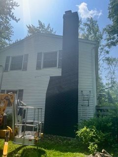 Two-story white house with a large black chimney and a tall black panel on the side, seen in a sunny yard