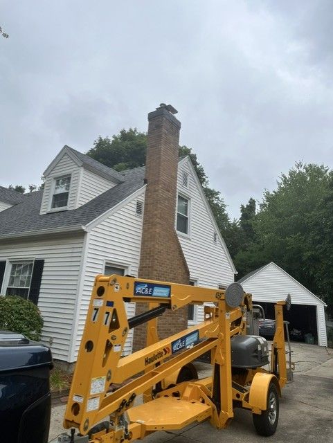 Yellow construction lift beside a white house with a brick chimney and garage under a cloudy sky