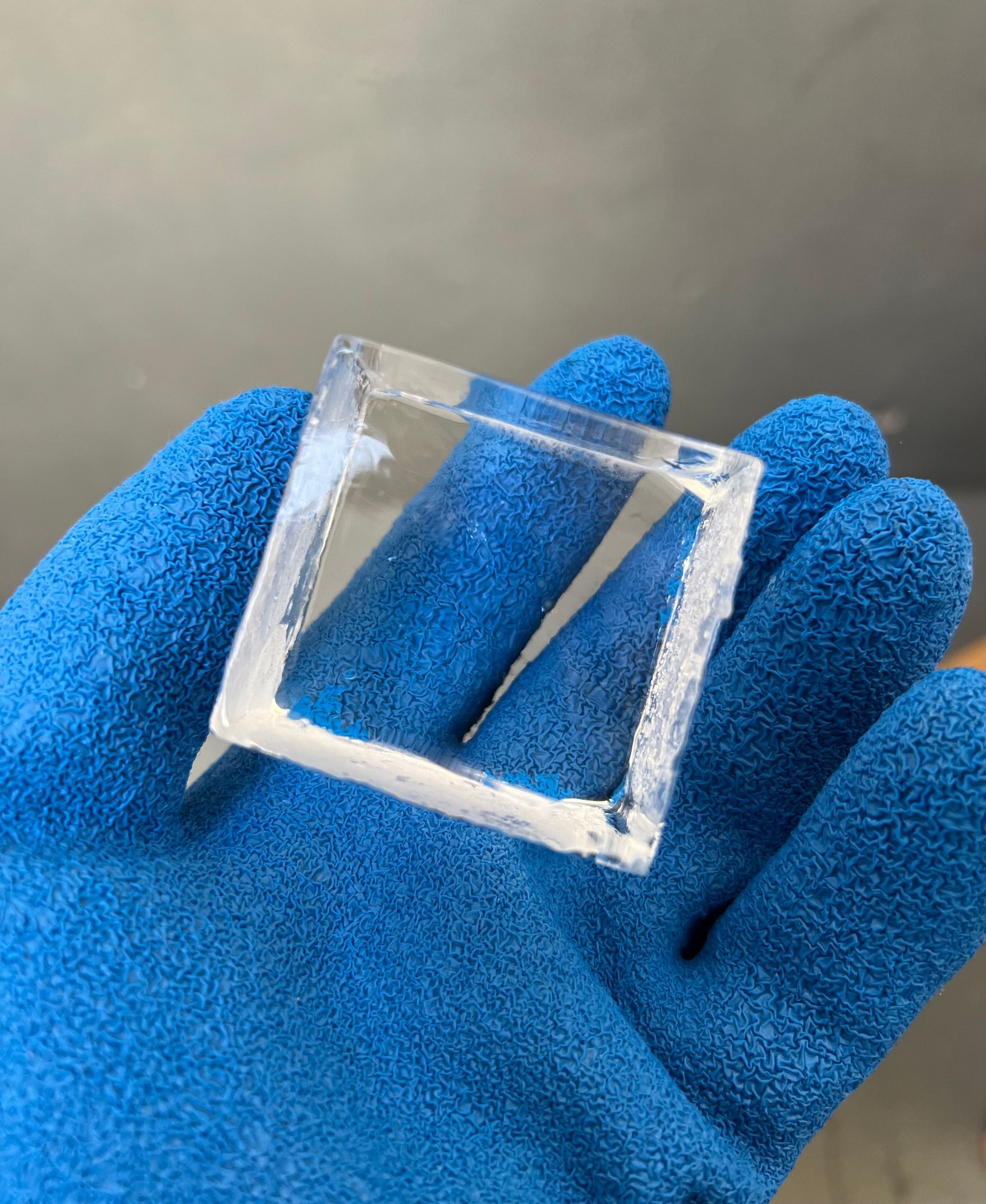 Blue-gloved hand holding a clear square plastic frame on a gray background