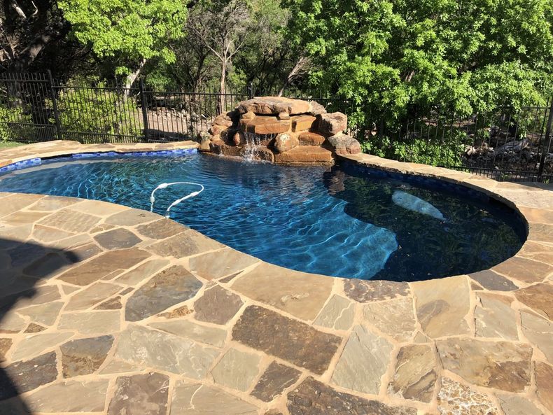 Curved backyard swimming pool with blue water, stone deck, and rock waterfall surrounded by trees