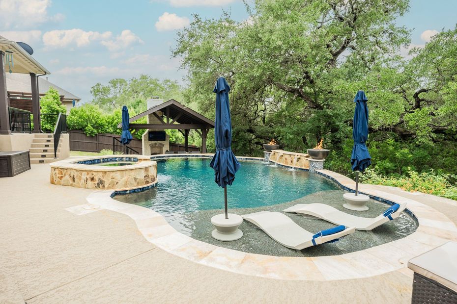 Backyard pool with blue umbrellas, lounge chairs, and a stone hot tub under trees