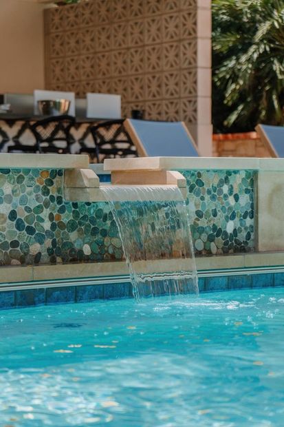 Pool waterfall with turquoise mosaic tiles beside lounge chairs and a patterned wall.