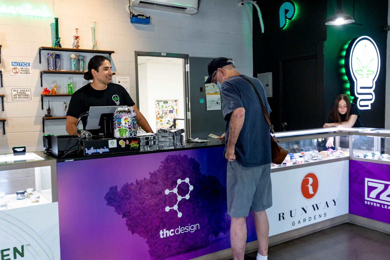 A customer at a dispensary counter with a cashier. The store interior has glass cases and colorful lighting.