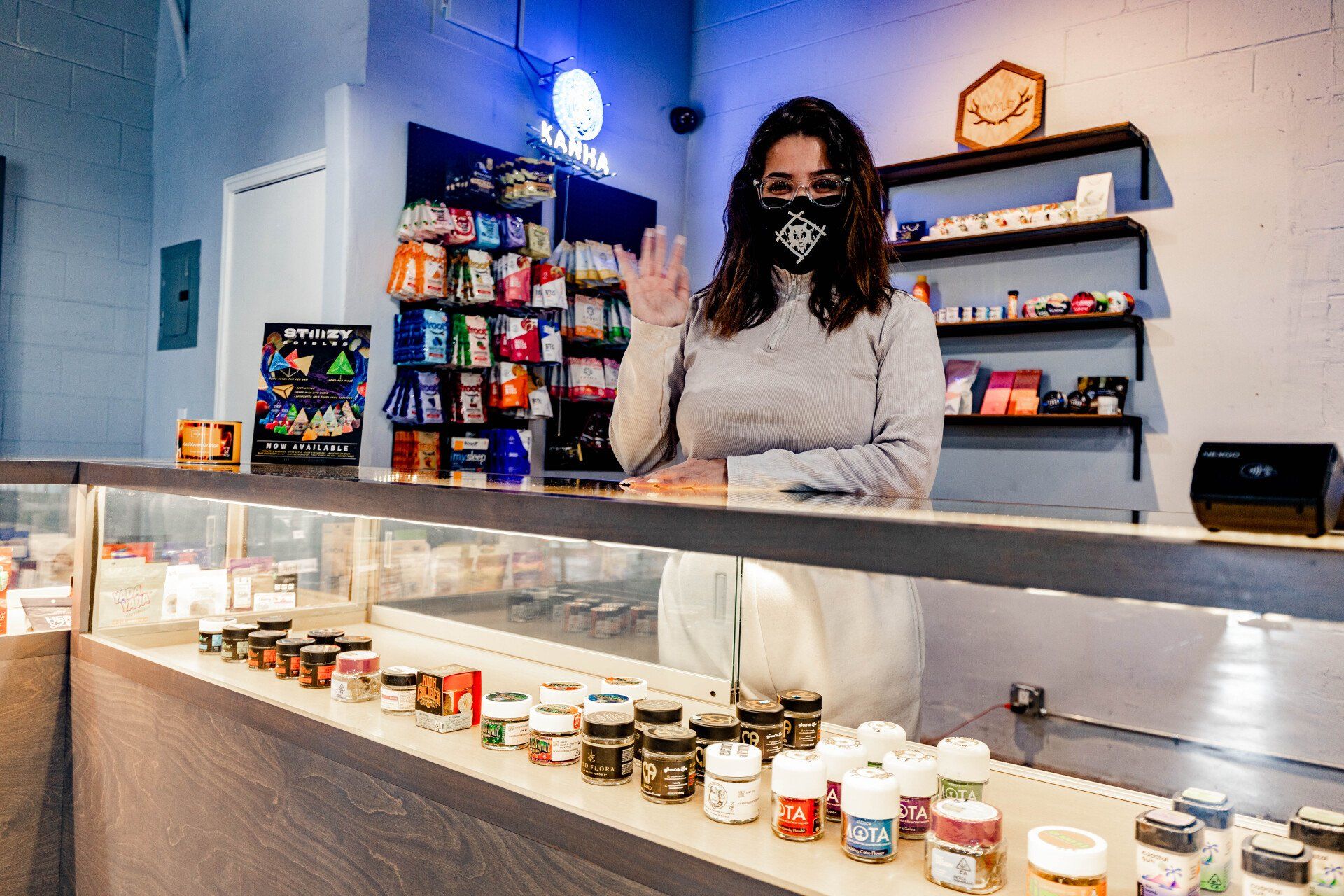 A woman wearing a mask waves from behind a dispensary counter filled with cannabis products.