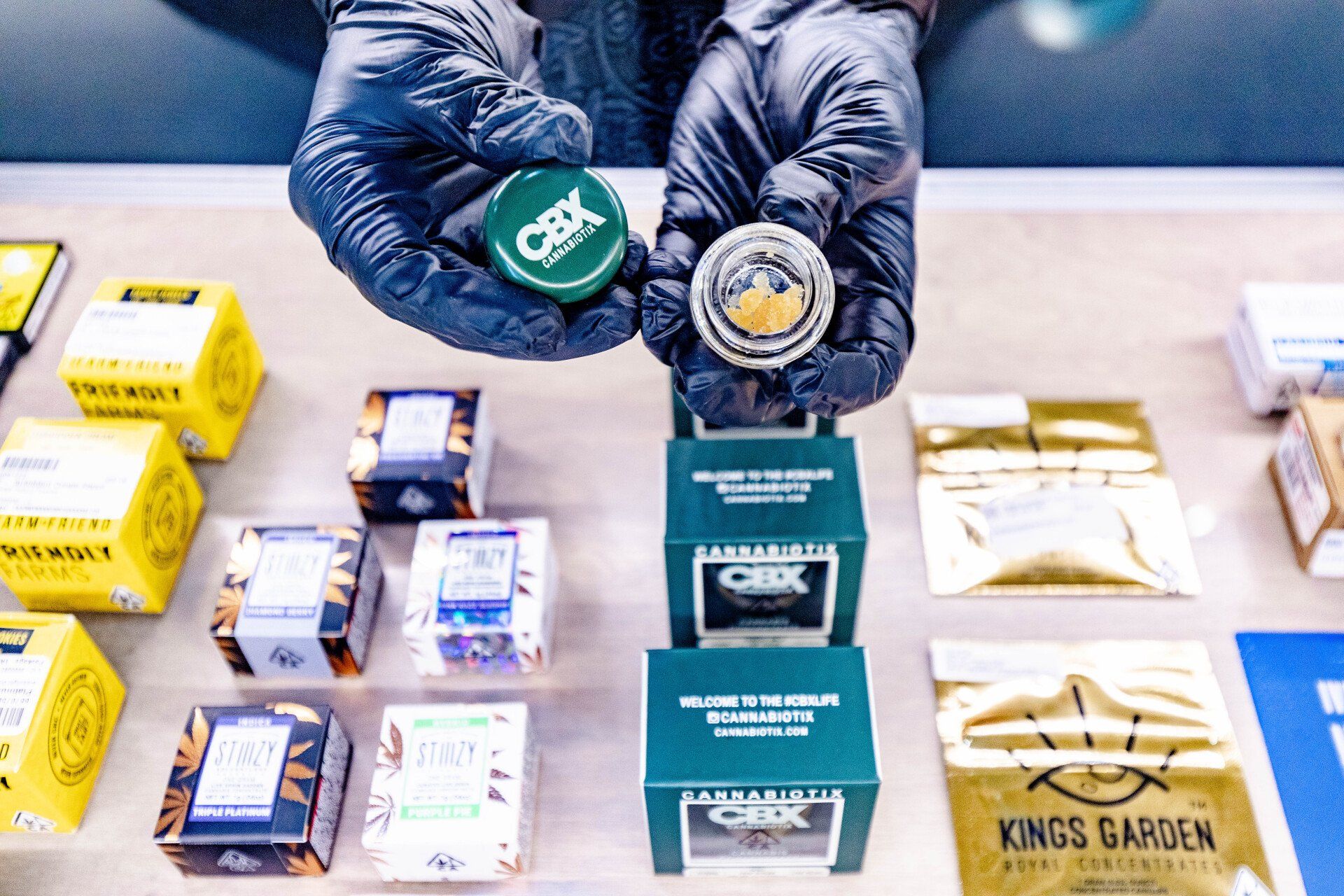 Hands in black gloves holding open cannabis container above display of packaged products.