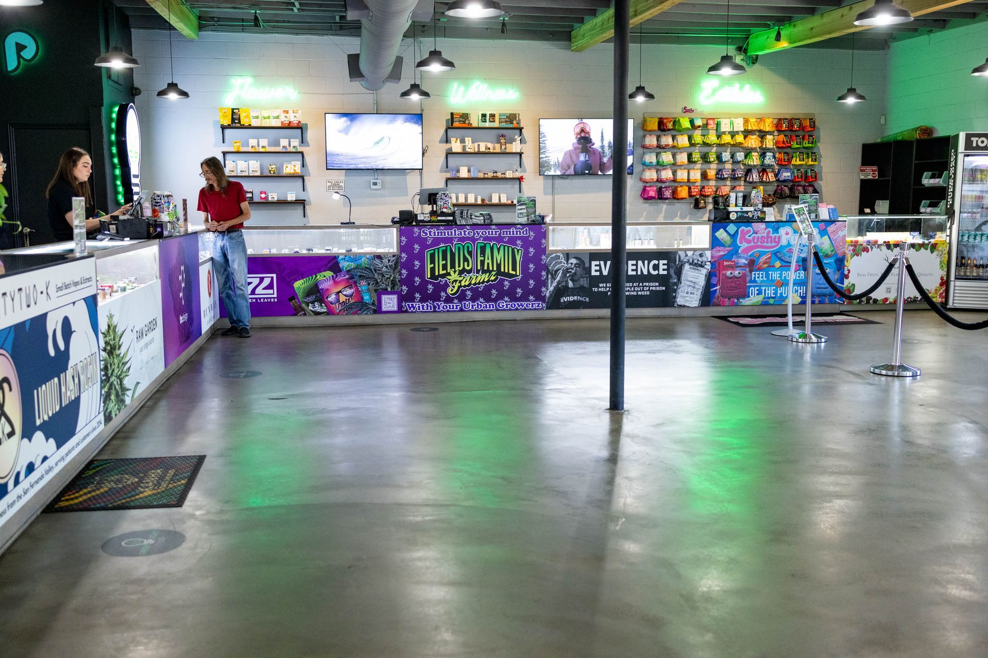 Inside a brightly lit dispensary with products on display. Two employees behind counters, one customer. Concrete floor, green and purple accents.