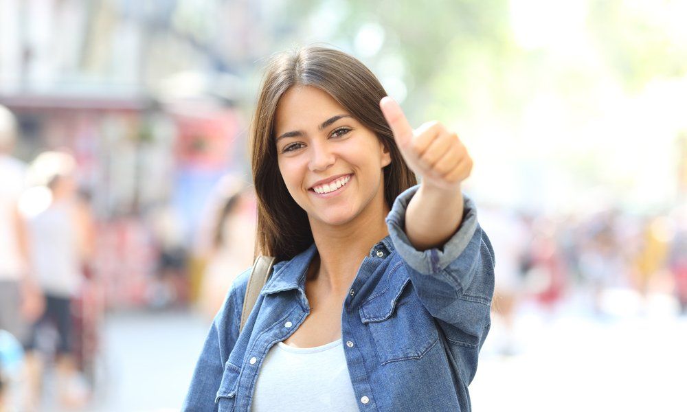 Woman in denim shirt smiles and gives thumbs up in a crowd outdoors.