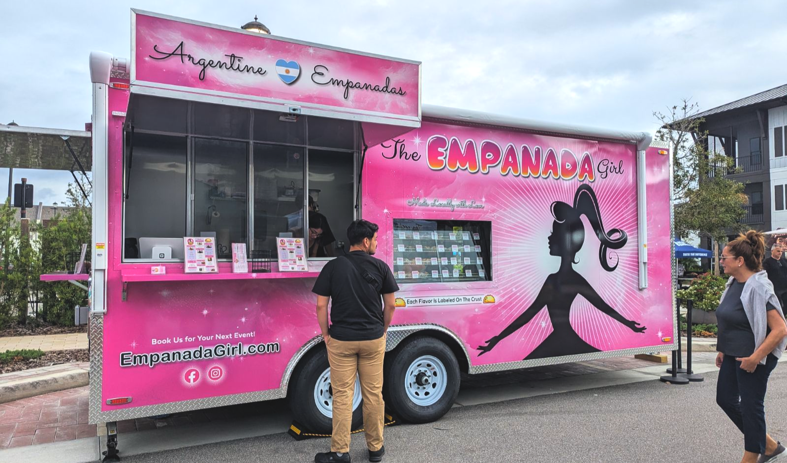 Pink empanada food truck with a serving window, menu signs, and two people standing nearby