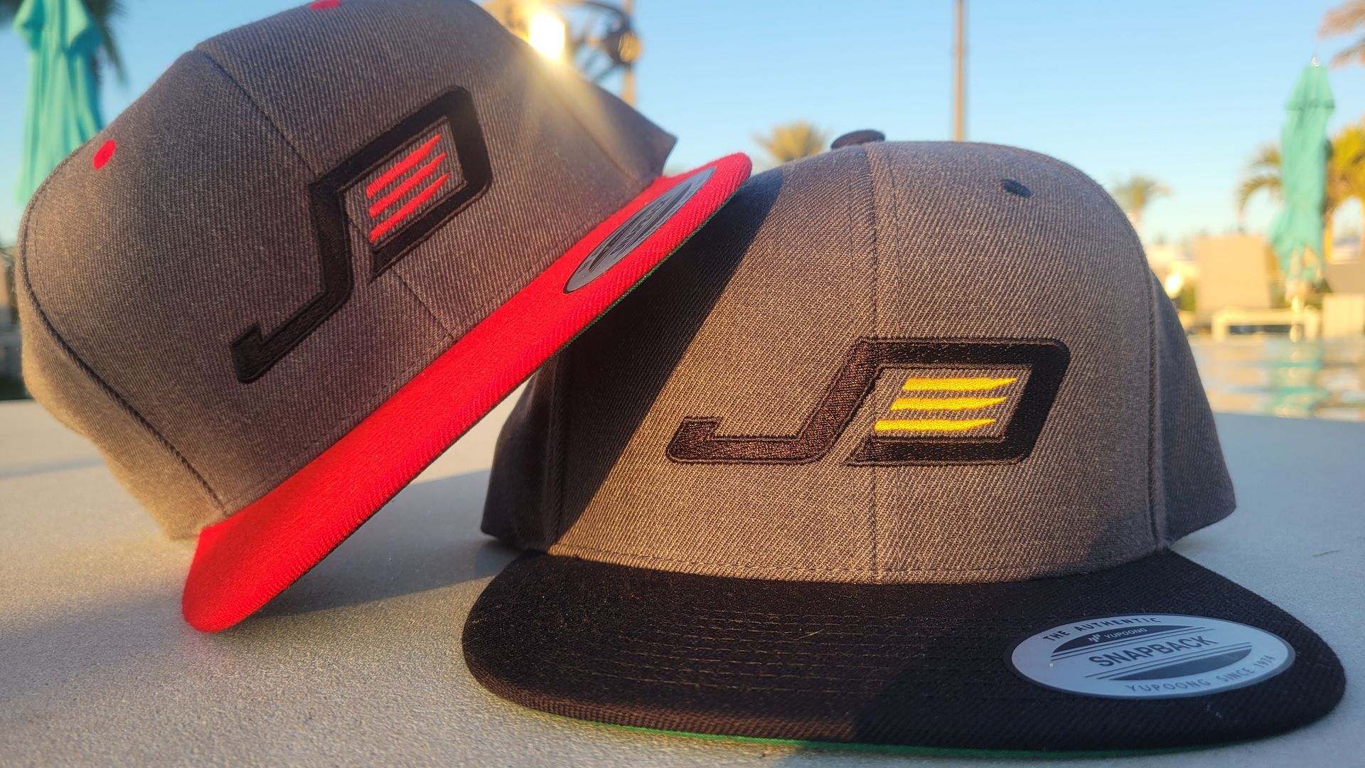 Two gray snapback hats with embroidered logo, one with red brim, other with black, outdoors.