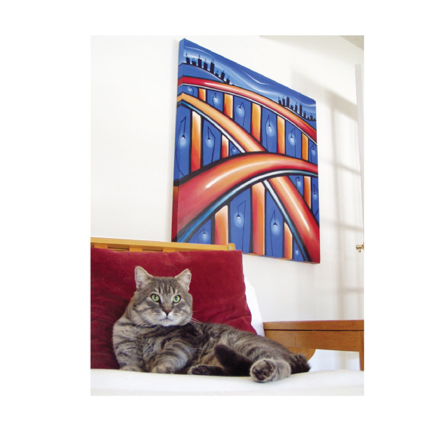 A tabby cat rests on a white chair with a red cushion, positioned below a large, colorful abstract painting of a bridge.