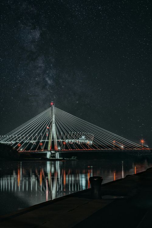 Nighttime cable-stayed bridge lit white over calm water, reflecting under a starry sky.