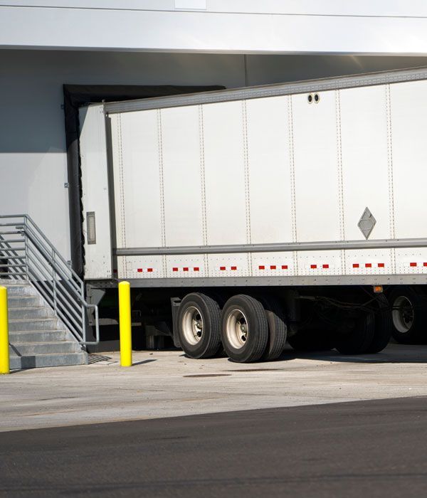 White semi-truck backed up to loading dock. Stairs, yellow posts, and grey building in view.