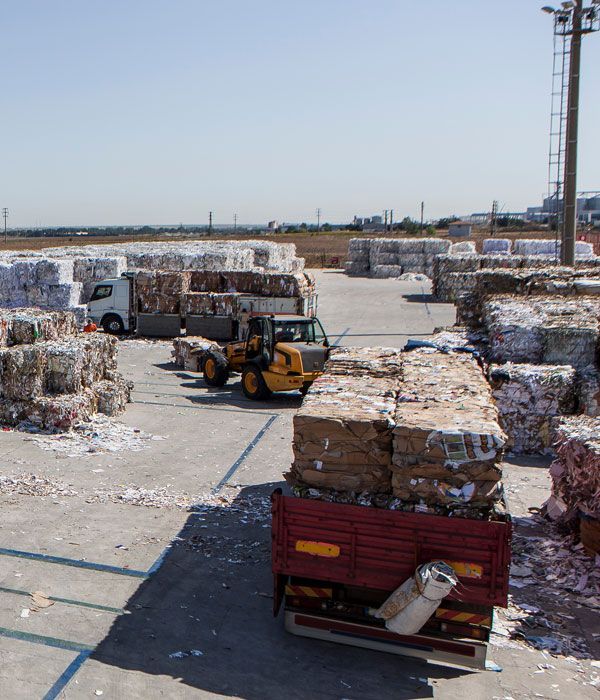Forklift loading a truck with stacked bales of recycled paper in an outdoor recycling facility.