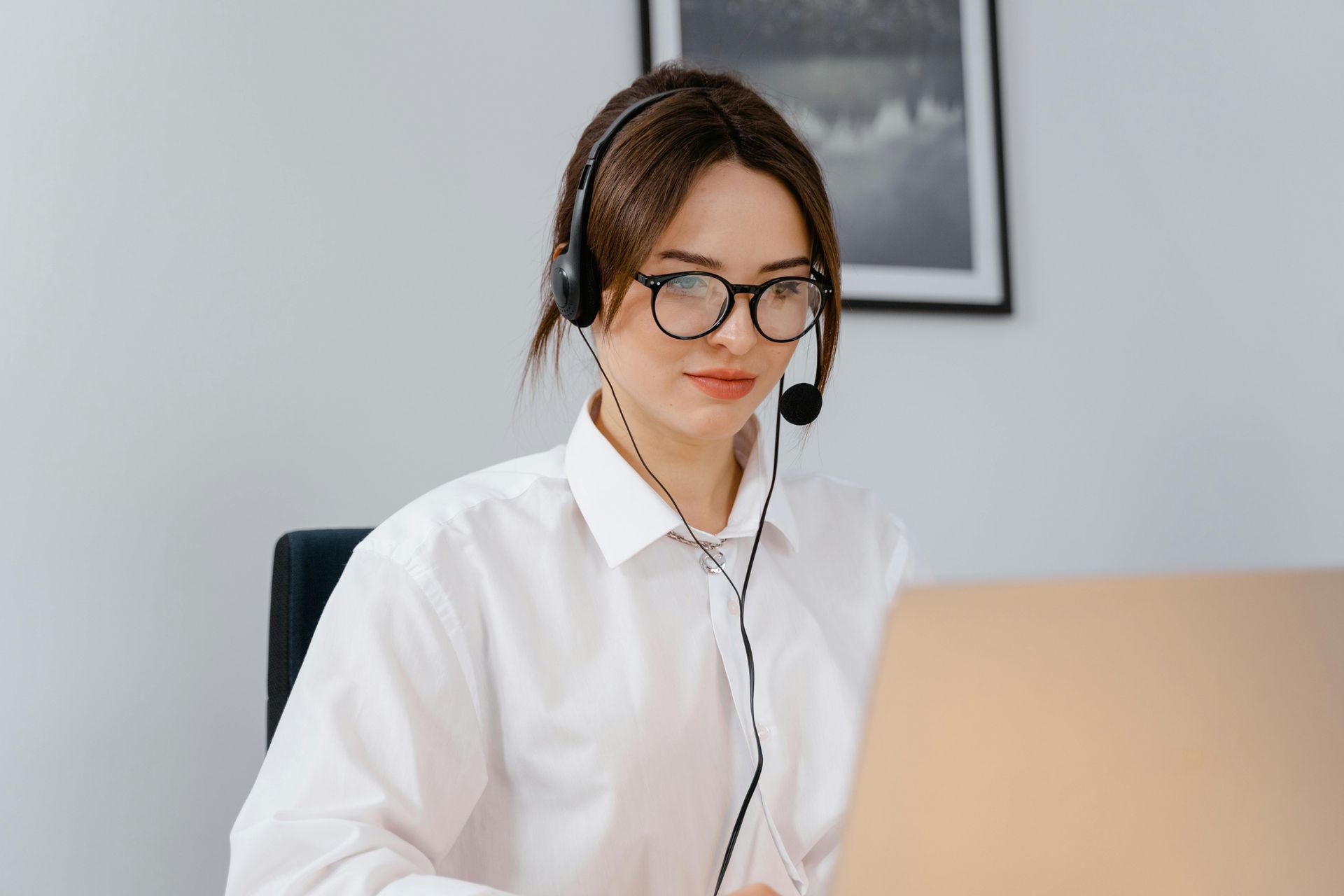 Woman wearing a headset and glasses, working on a laptop in a light-colored office.