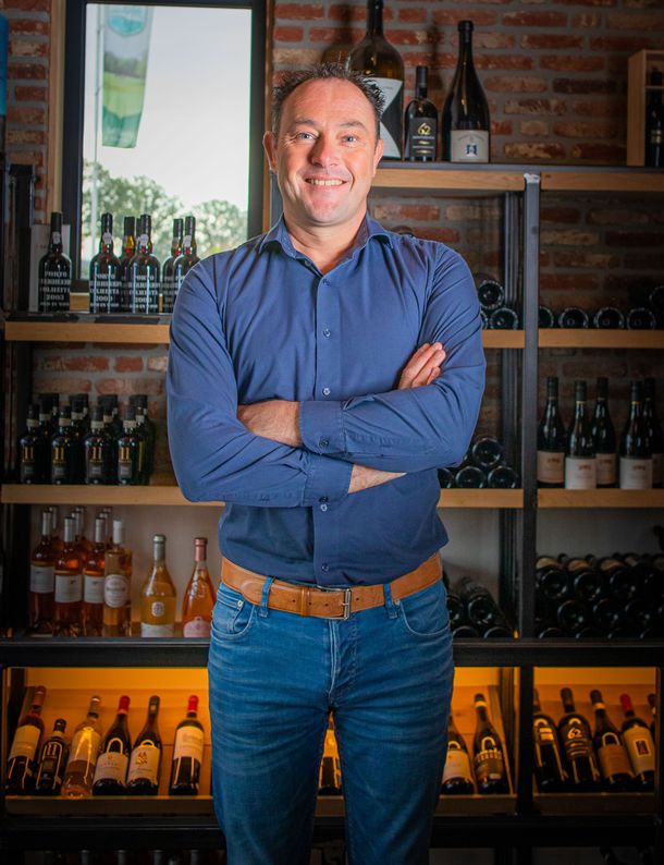 Een lachende man in een blauw shirt en spijkerbroek staat met zijn armen over elkaar in een wijnwinkel.