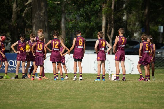 Maroon-and-gold athletes huddle on a grassy field, seen from behind near the sideline.