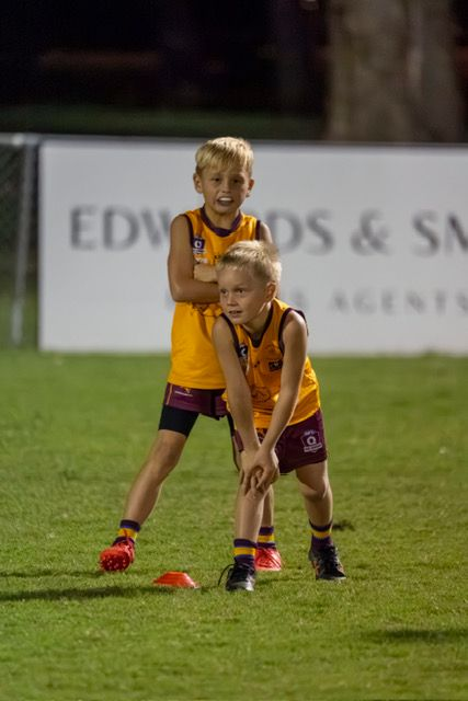 Two young players in yellow jerseys on a lit soccer field, one bending over red cones.