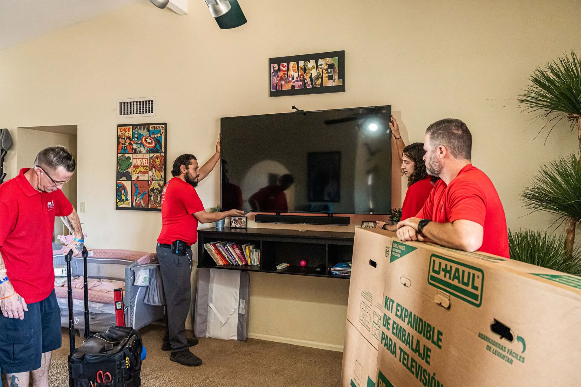Three people in red shirts move a large TV during a room move, with a U-Haul box in the foreground.