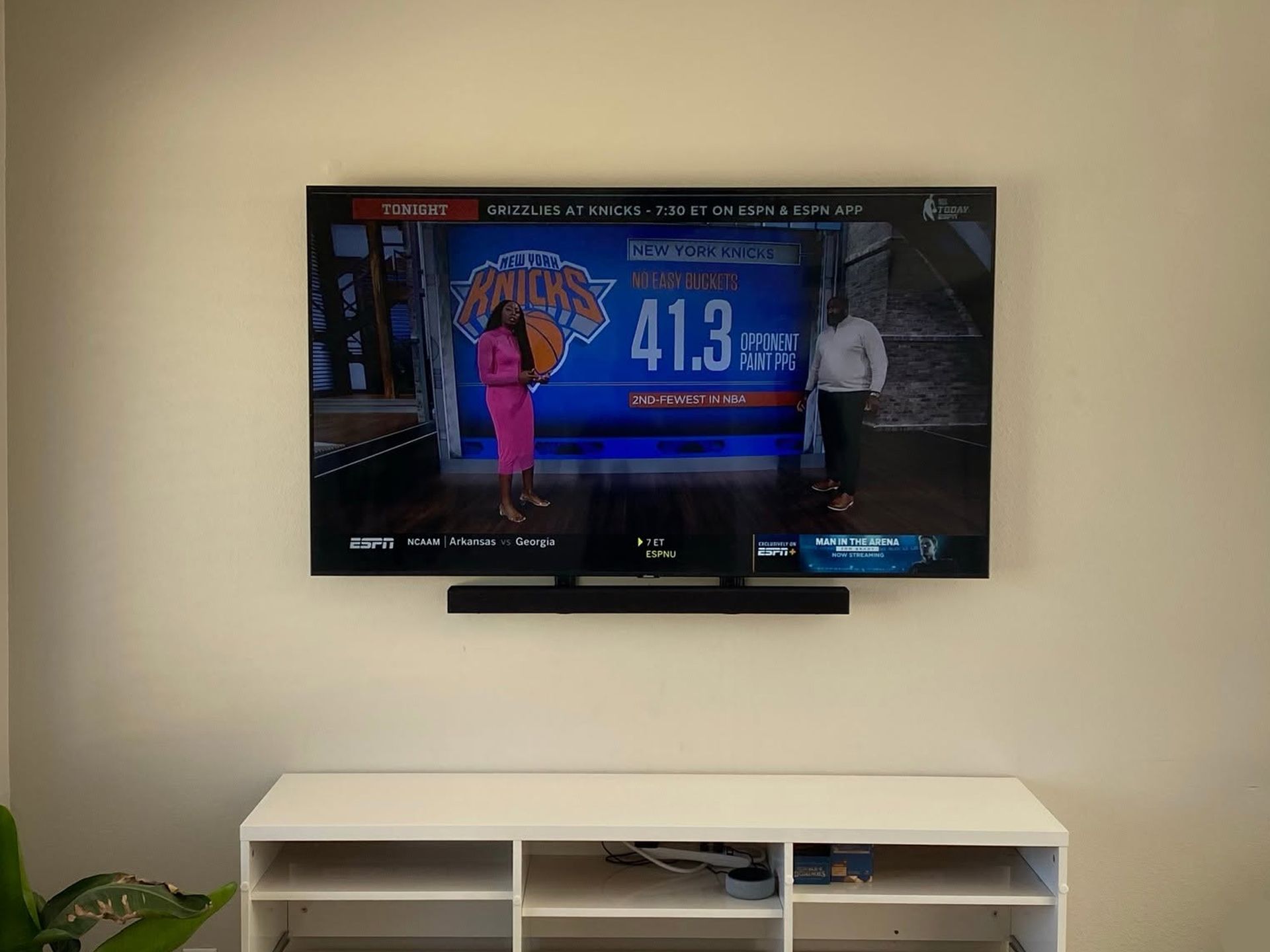 Wall-mounted TV showing a weather broadcast above a white media console in a beige room