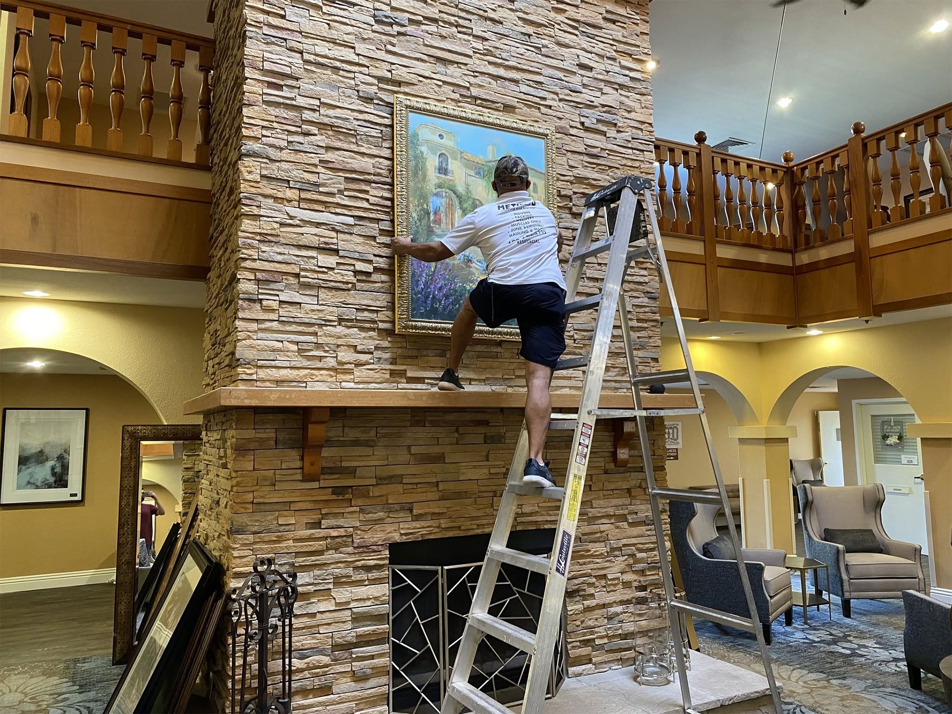 Person on a ladder hanging a painting on a stone fireplace in a lodge-style room
