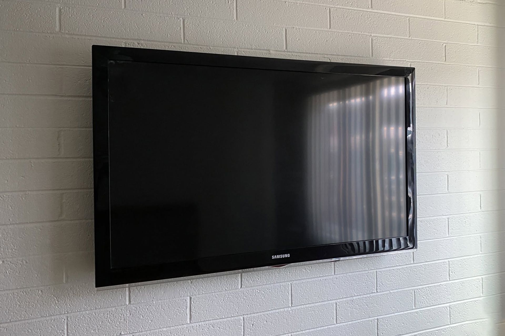 Black flat-screen TV mounted on a white brick wall, reflecting light from a window.