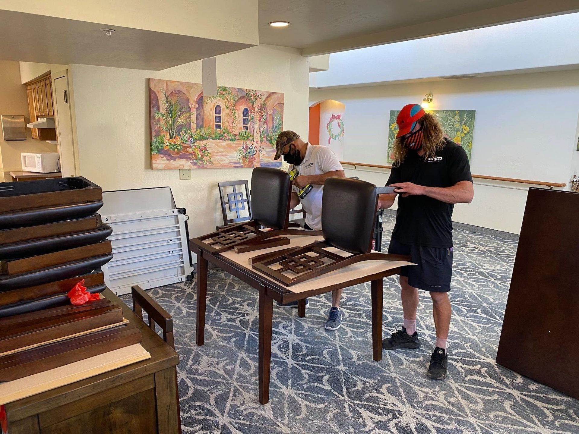 Two people assembling tables in a bright room with stacked chairs and colorful wall art.