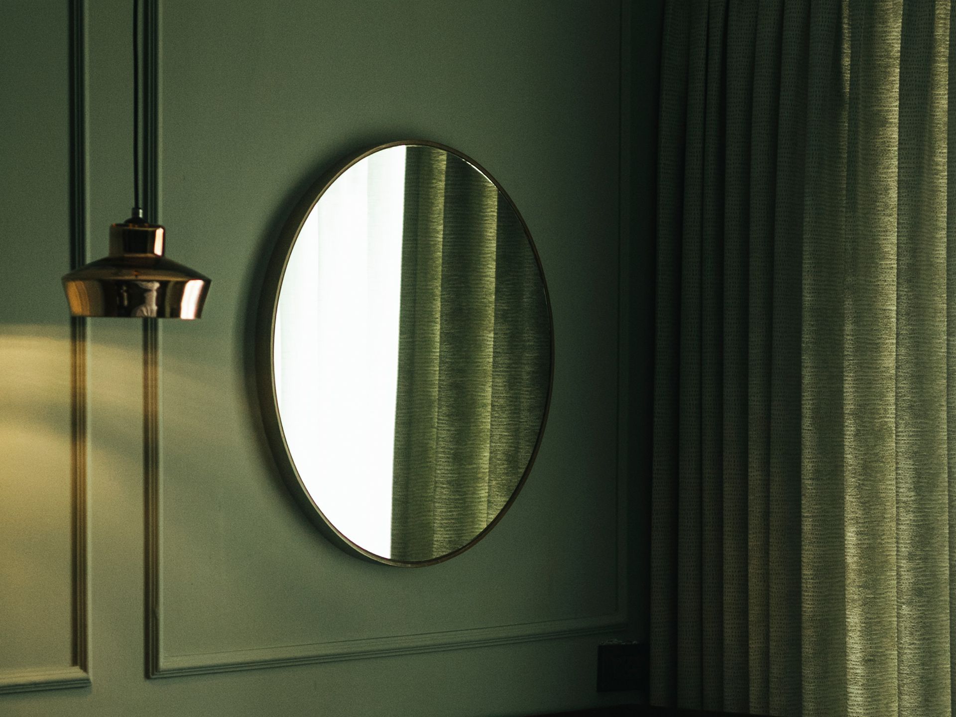 Oval mirror on a dark green paneled wall beside a hanging lamp and curtain.