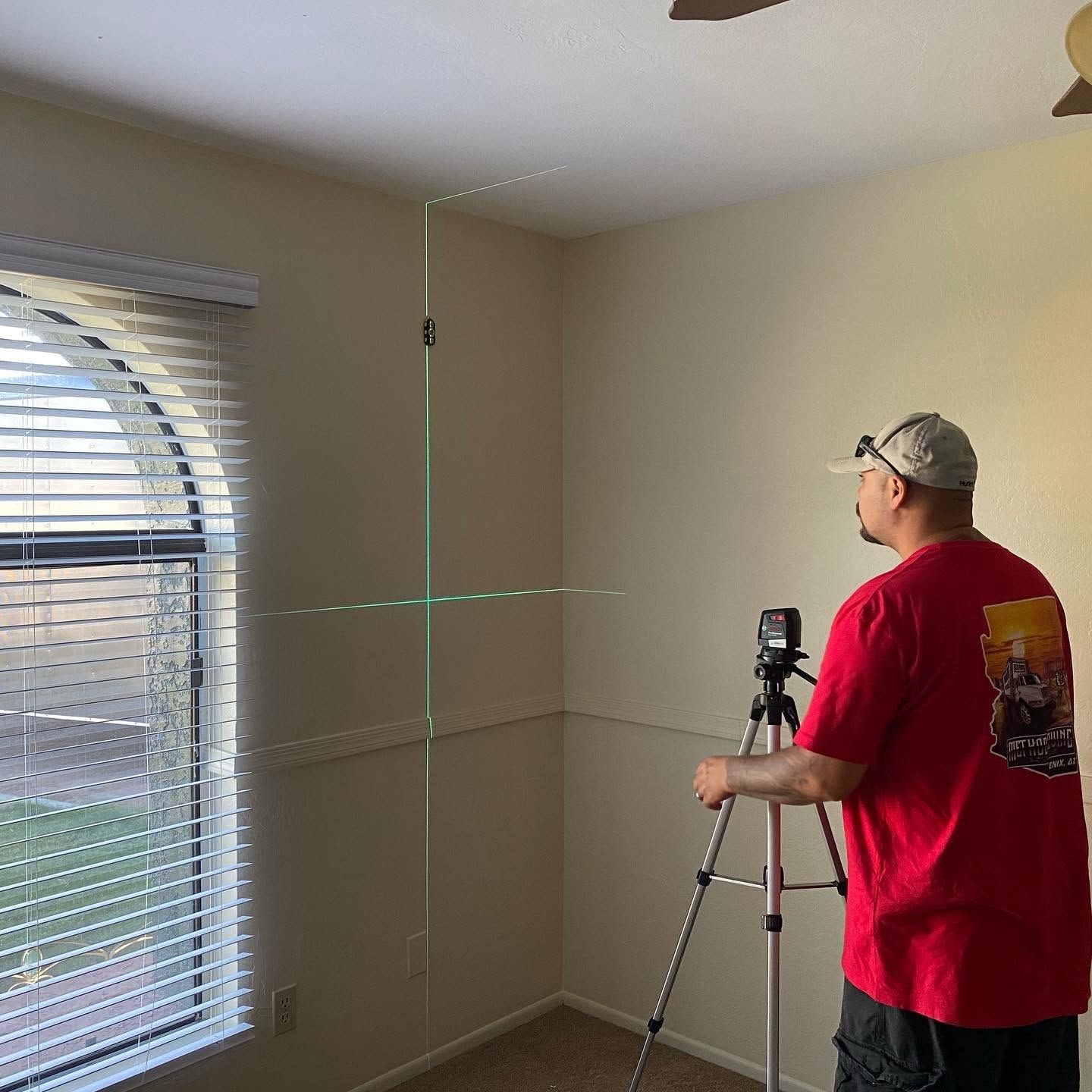 Person in red shirt using a tripod-mounted laser level to mark lines on a beige room wall near a window.