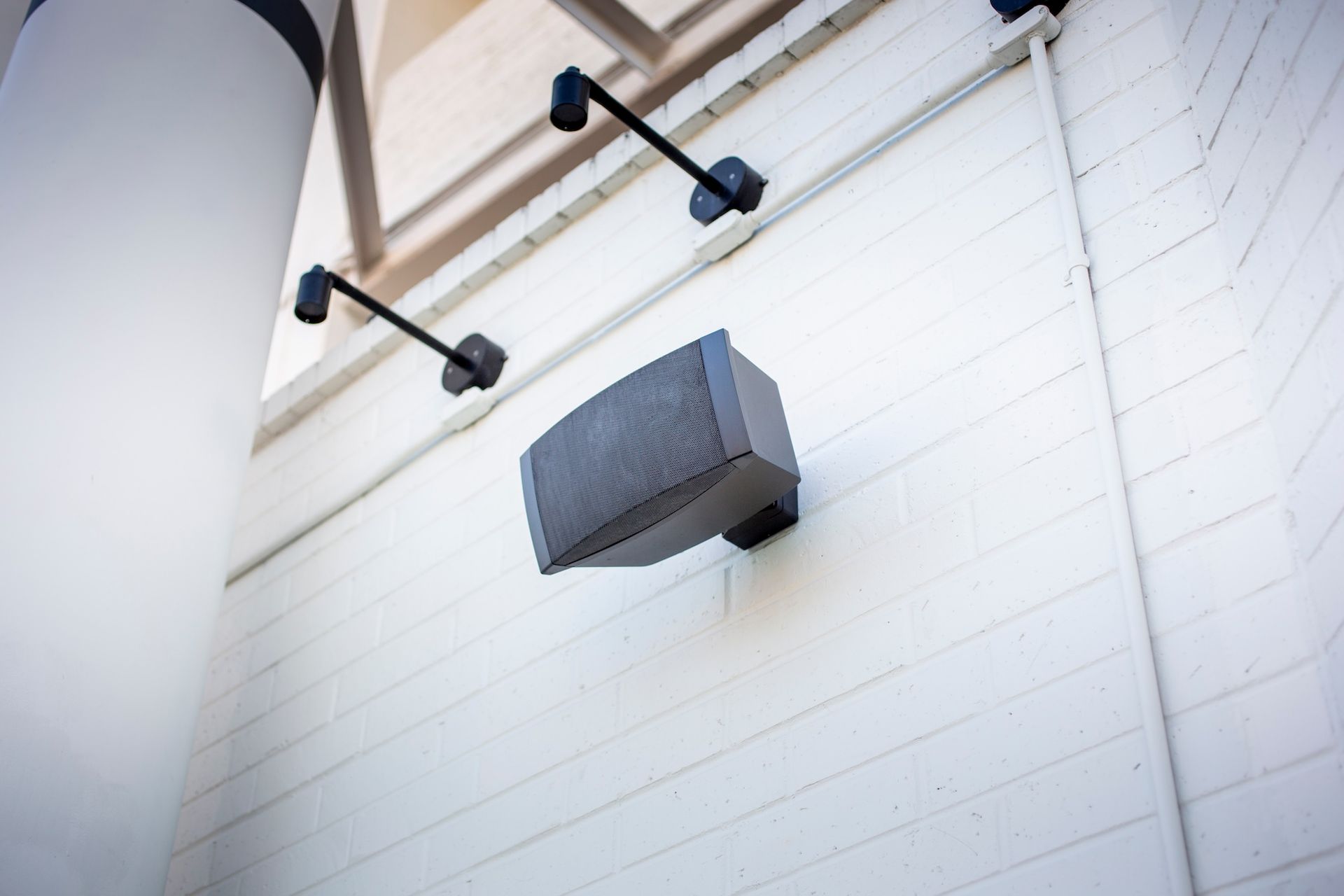 White exterior wall with a gray speaker mounted below black metal brackets near the ceiling.