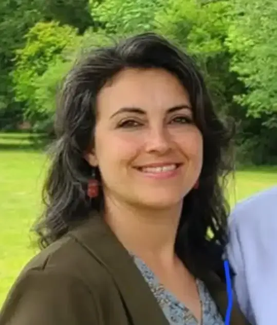 Woman with dark hair smiles outdoors, wearing a green jacket and patterned shirt.