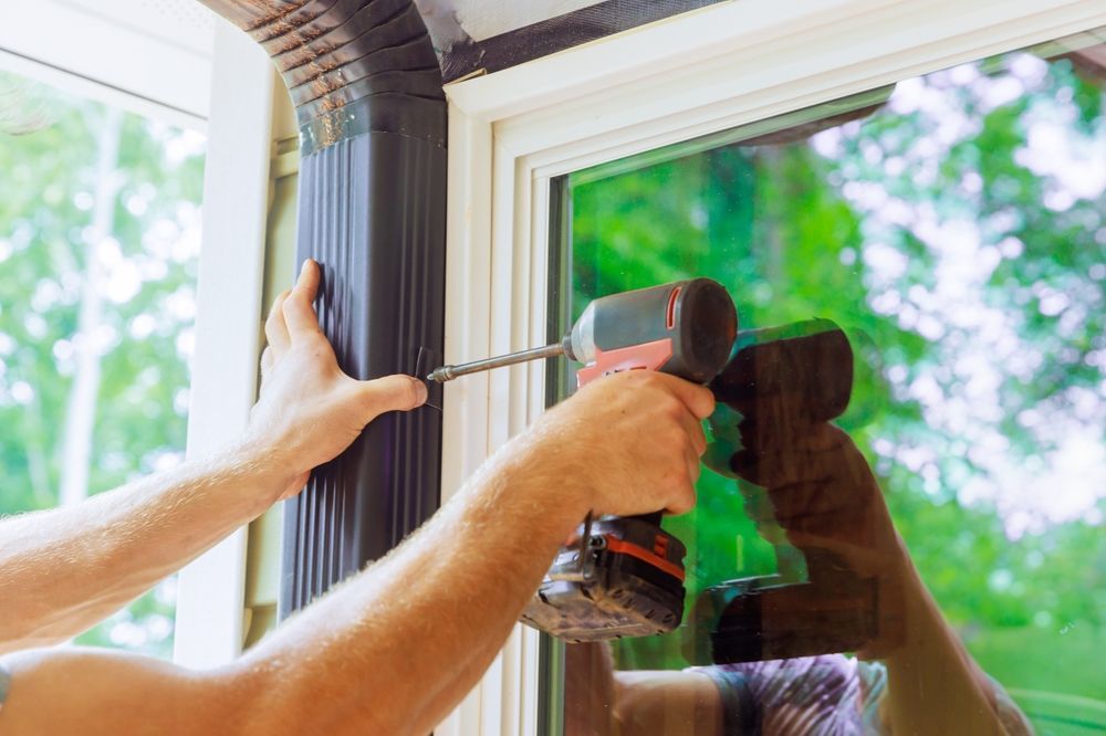 Hands installing a black window frame with a drill beside a bright green garden view