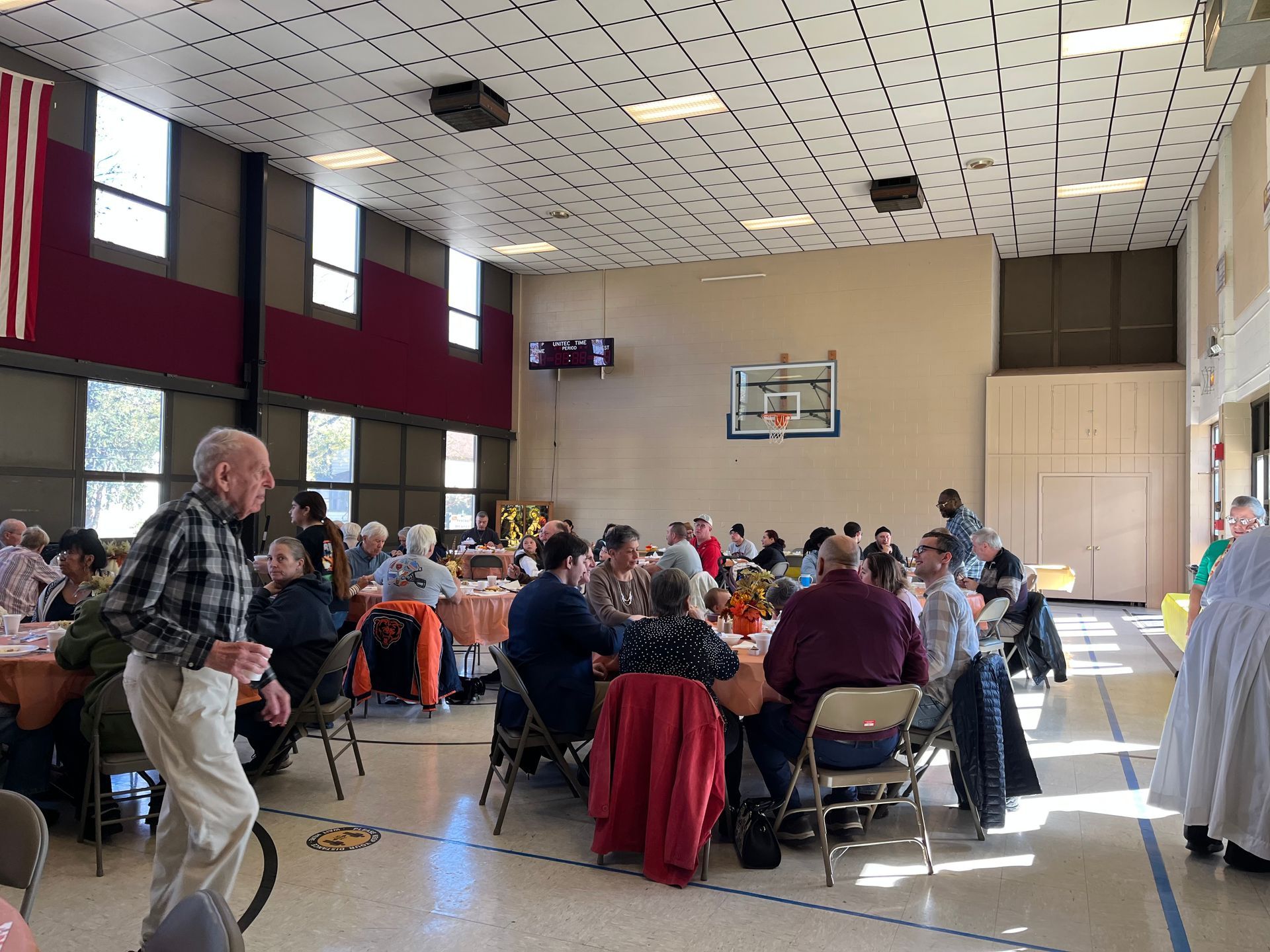 Community dining hall with people seated at tables during a gathering, with a man standing near the left side.