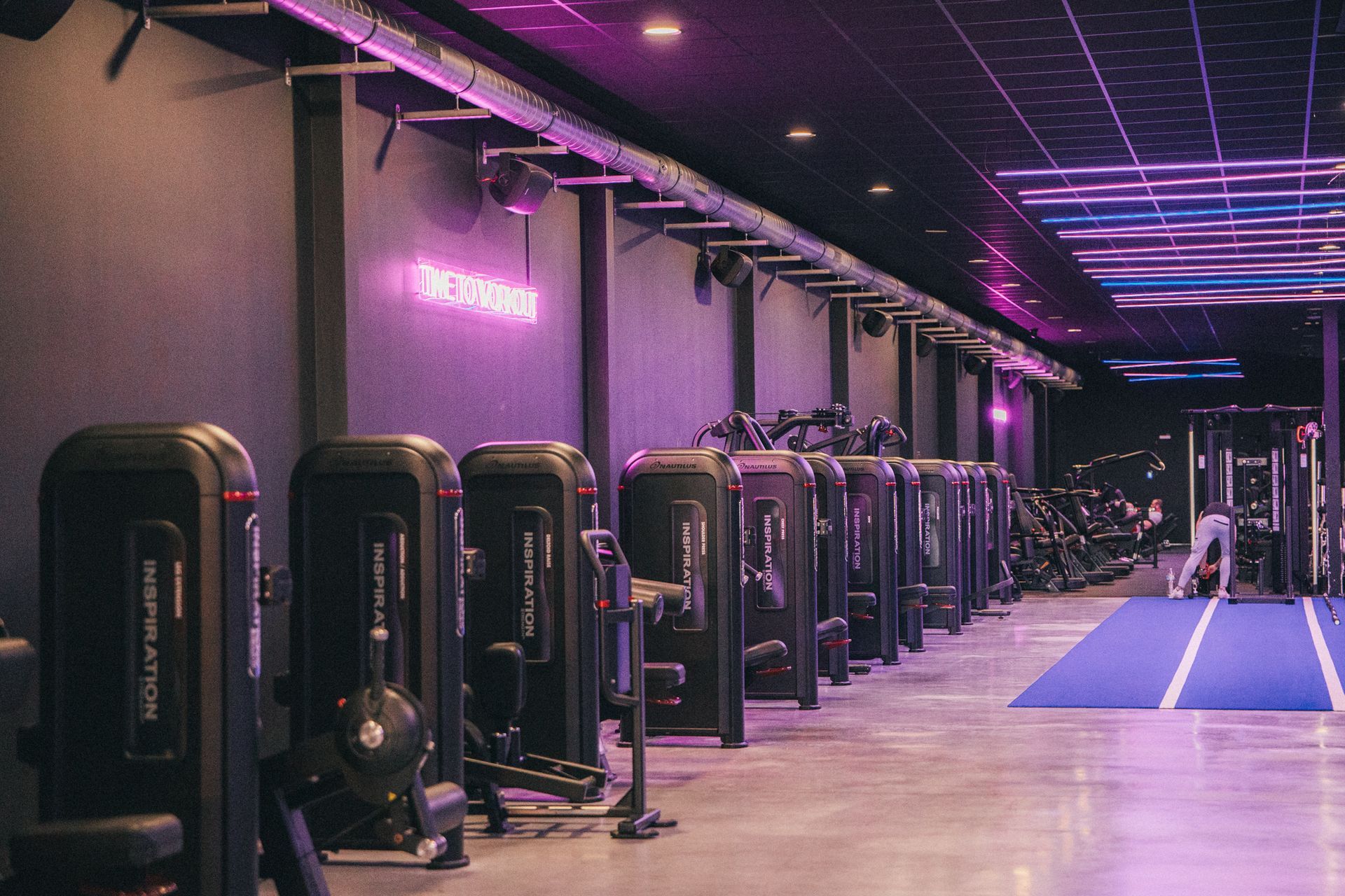 Een grote sportschool vol met fitnessapparatuur en blauwe verlichting.