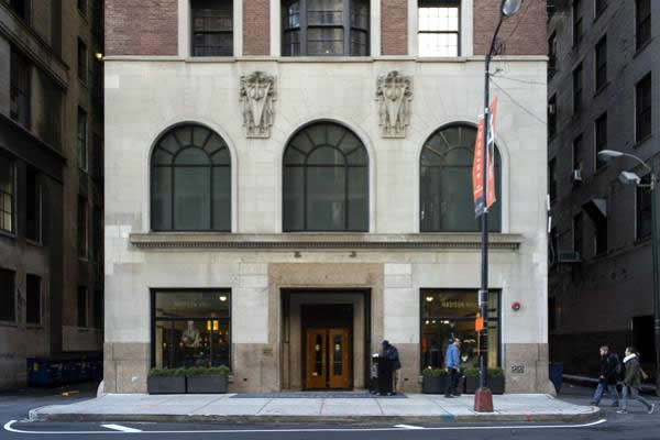 Building facade with arched windows, light stone, and brick. People stand outside the entrance.