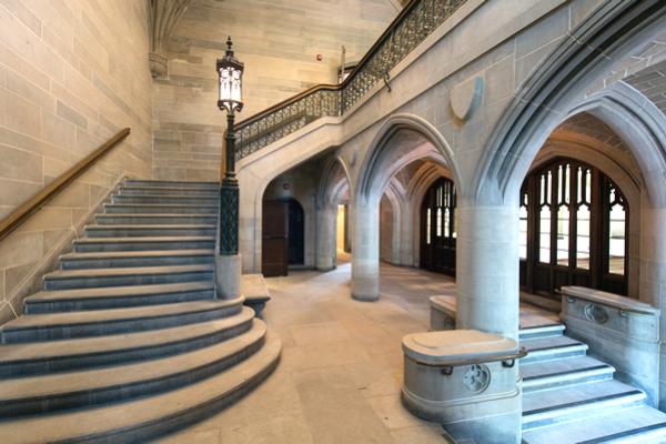 Stone staircase and arched hallway in a Gothic-style building, featuring a decorative lamp and stained-glass windows.