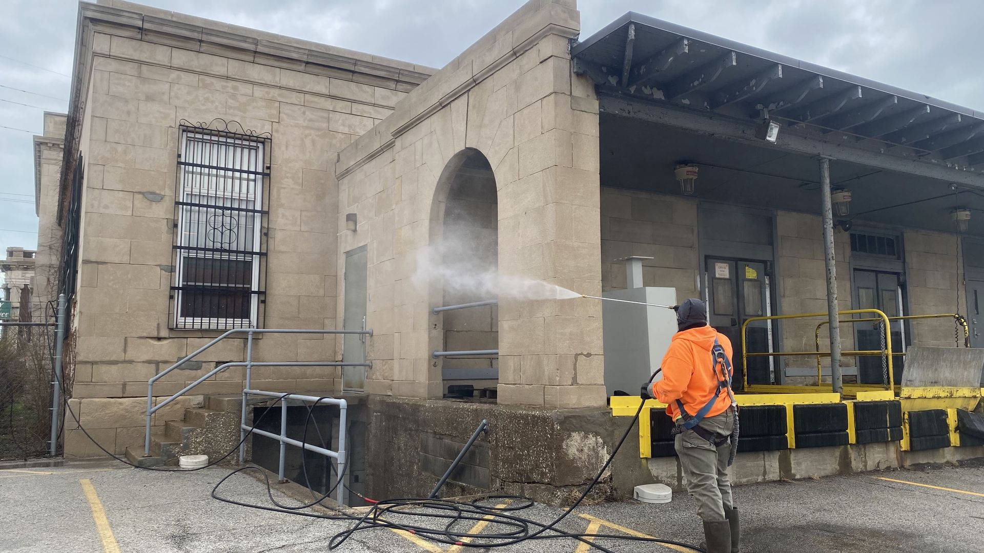 A RestoreWorks employee in an orange high-visibility hoodie and safety harness pressure washes a historic limestone building. A fine mist sprays from the nozzle as they clean the stone surface, removing dirt and buildup. The worker stands on a wet pavement near a loading dock, with hoses extending across the ground. The building shows signs of aging, including discoloration and minor damage to the masonry.