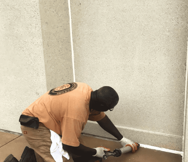 A RestoreWorks employee kneels while applying caulk along the base of a textured concrete wall. Wearing gloves and using a caulking gun, they carefully seal the joint to protect against water infiltration and structural wear. The worker is dressed in a company-branded shirt and equipped with a tool pouch for efficiency.