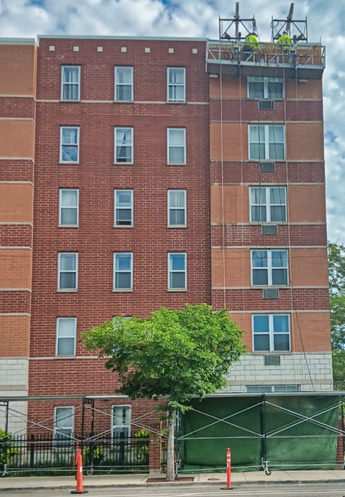 Brick apartment building with rooftop equipment, green tree, and fenced sidewalk in front