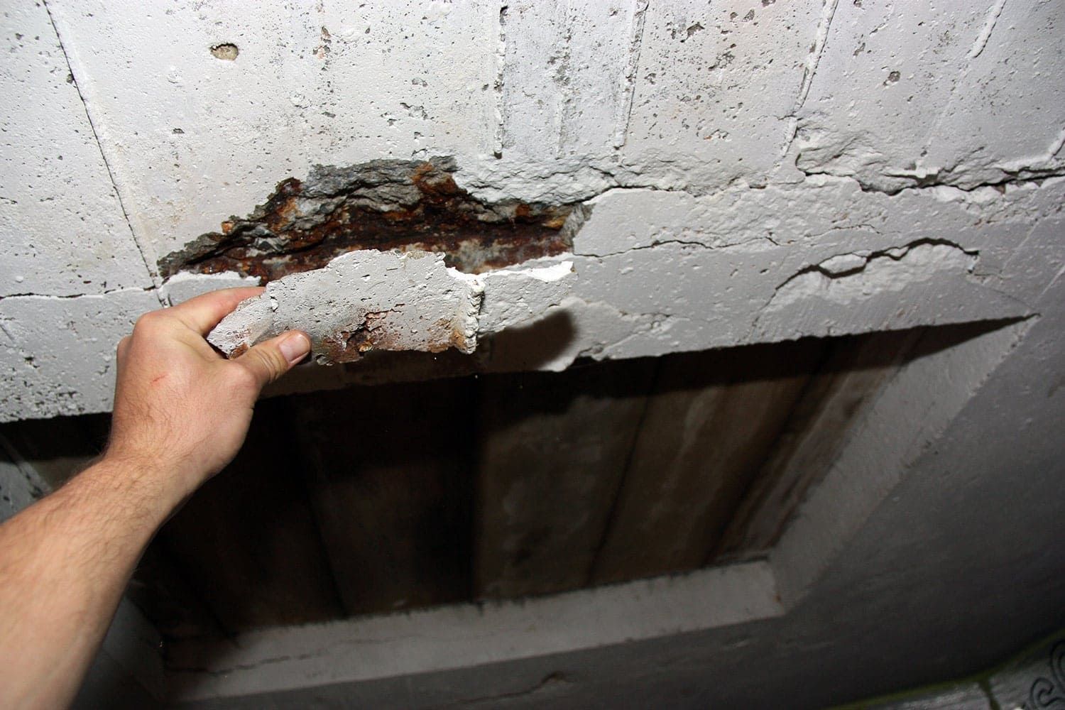 A hand peels a loose, crumbling section of concrete from a ceiling, revealing rusted metal reinforcement underneath.