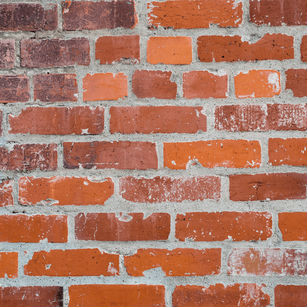 A close-up of a brick wall with deteriorating mortar joints. The mortar appears worn and eroded, with visible cracks and gaps between the bricks. Some bricks show signs of chipping and surface damage. This type of deterioration is often caused by winter weather, as freeze-thaw cycles weaken the mortar, leading to structural issues and increased water infiltration into the masonry wall.