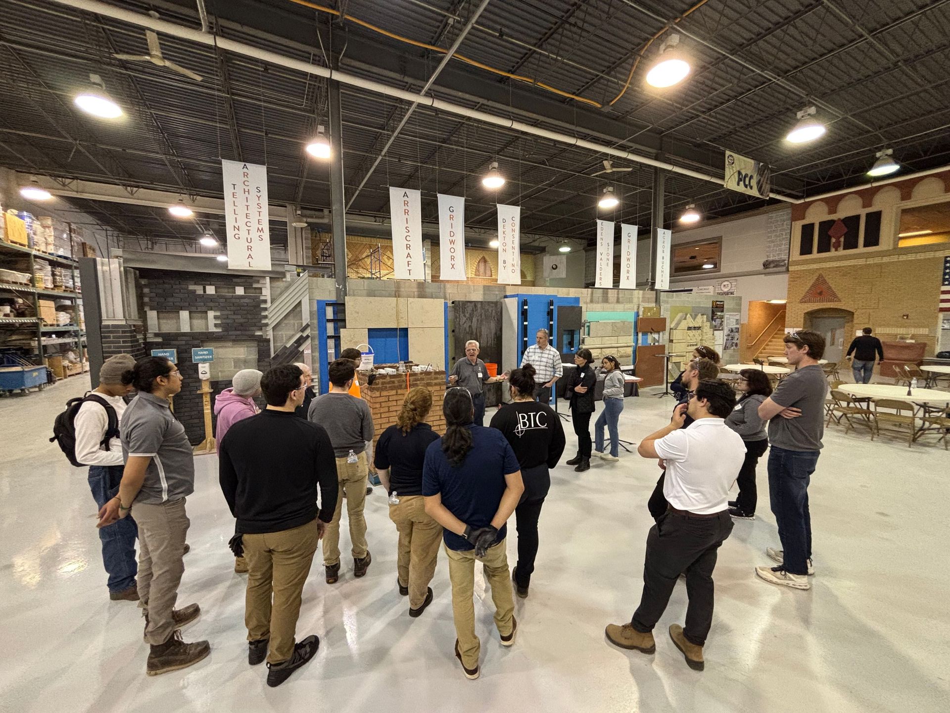 A group of people standing in a large warehouse space listening to a speaker standing near construction mock-ups.