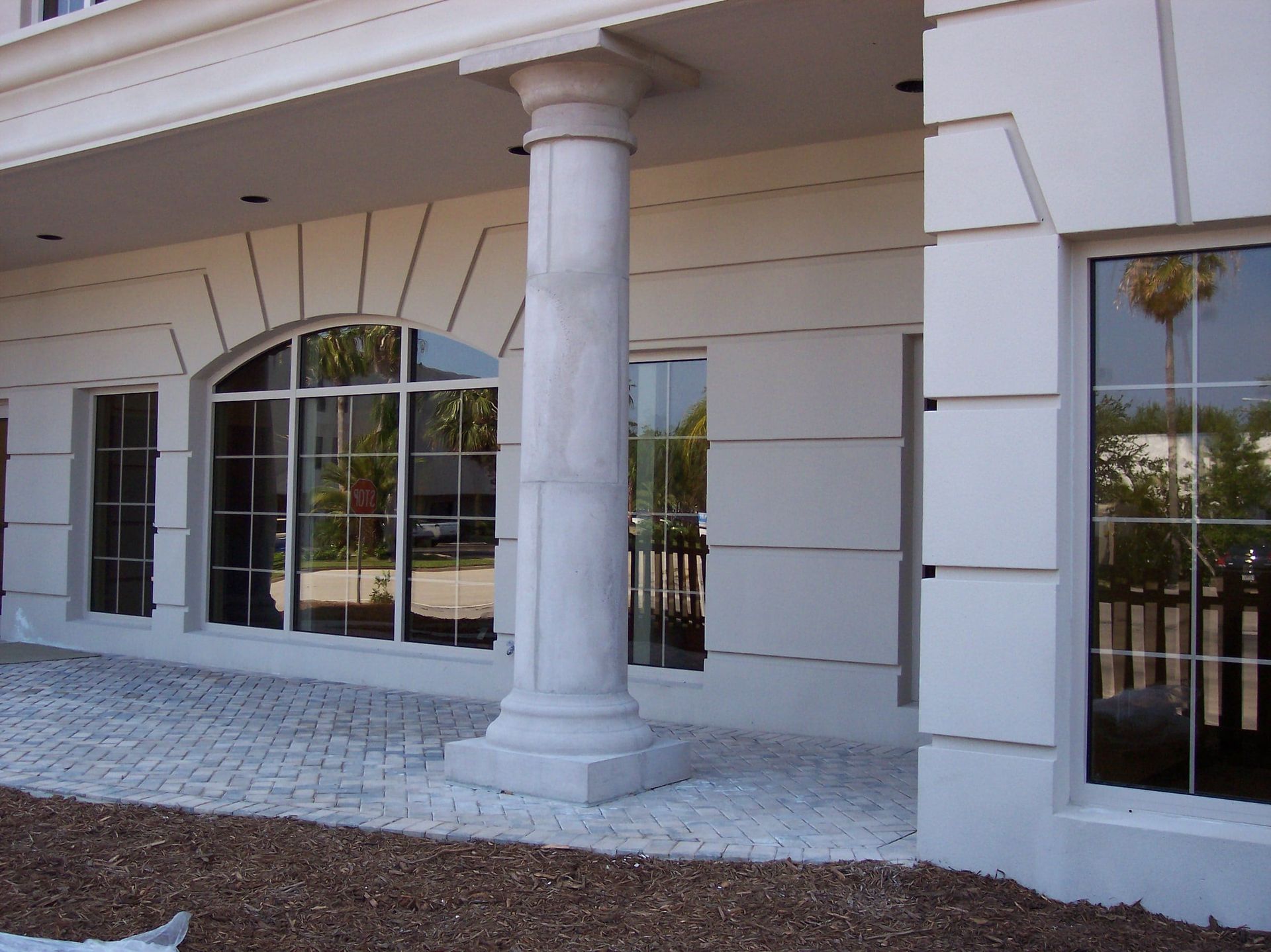 A white building facade with a large arched window, a central classical column, and a paved patio area.