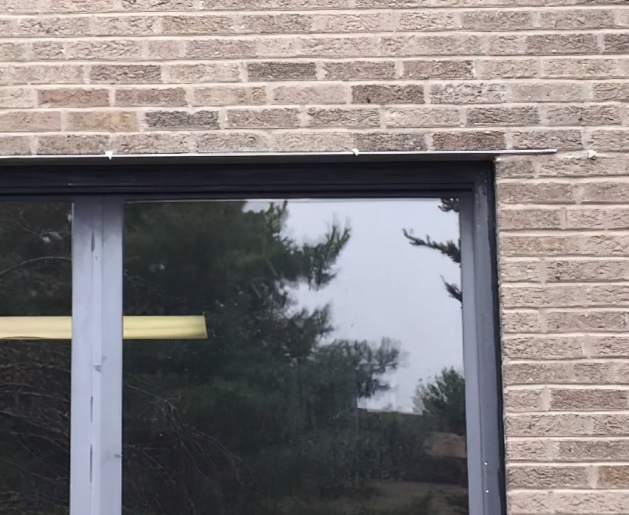 Brick wall above a window with dark frame. Reflective glass shows trees and sky.