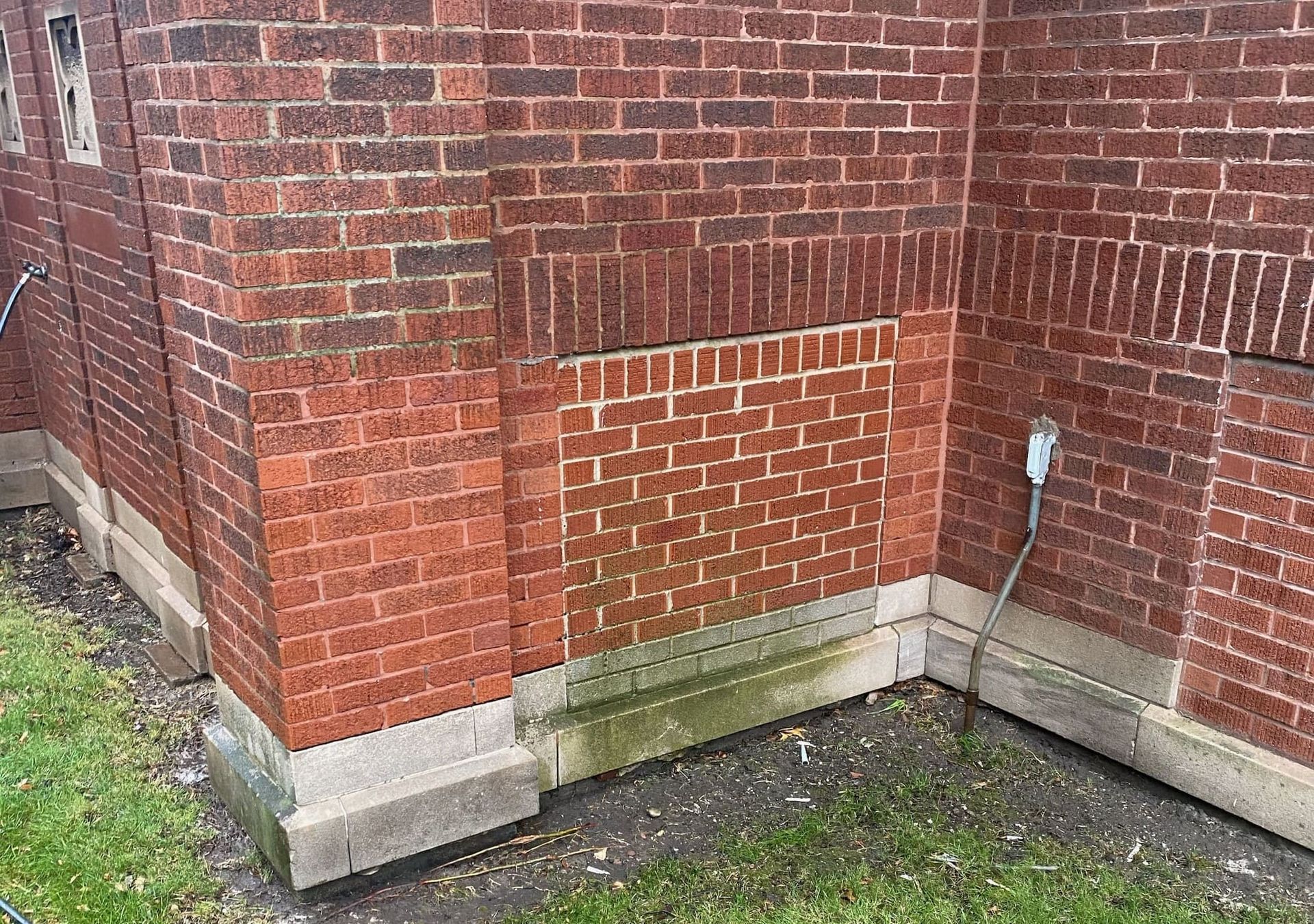 A corner of a red brick building with a stone foundation and a section of brick infill, viewed from an exterior lawn.