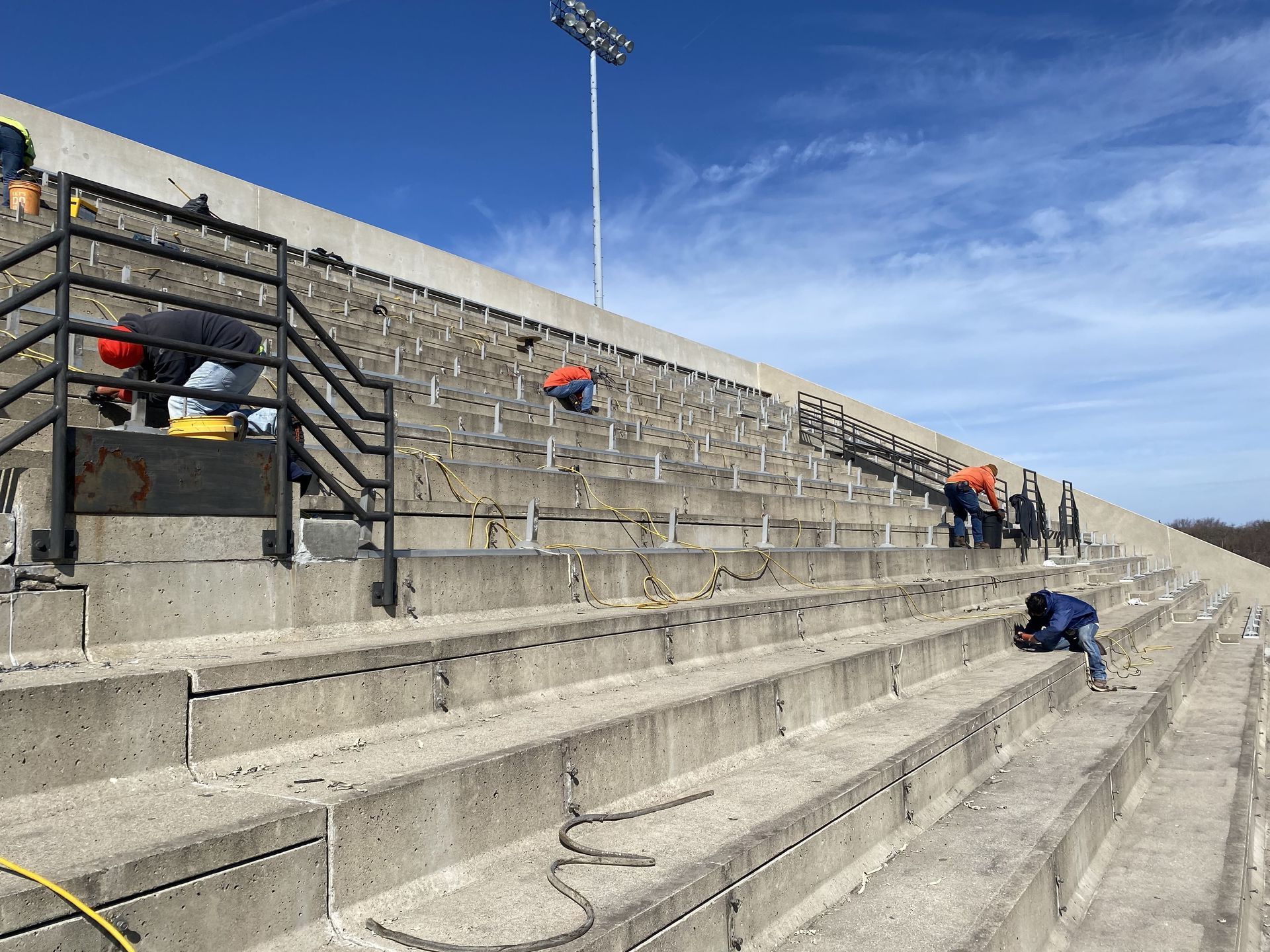 RestoreWorks crew members perform caulking and concrete restoration work at Merrillville High School's football stadium. Workers are spread across the concrete bleachers, sealing joints and repairing cracks to ensure the structure's longevity. Equipment, hoses, and buckets are visible throughout the site, while a bright blue sky provides a backdrop for the large outdoor stadium.