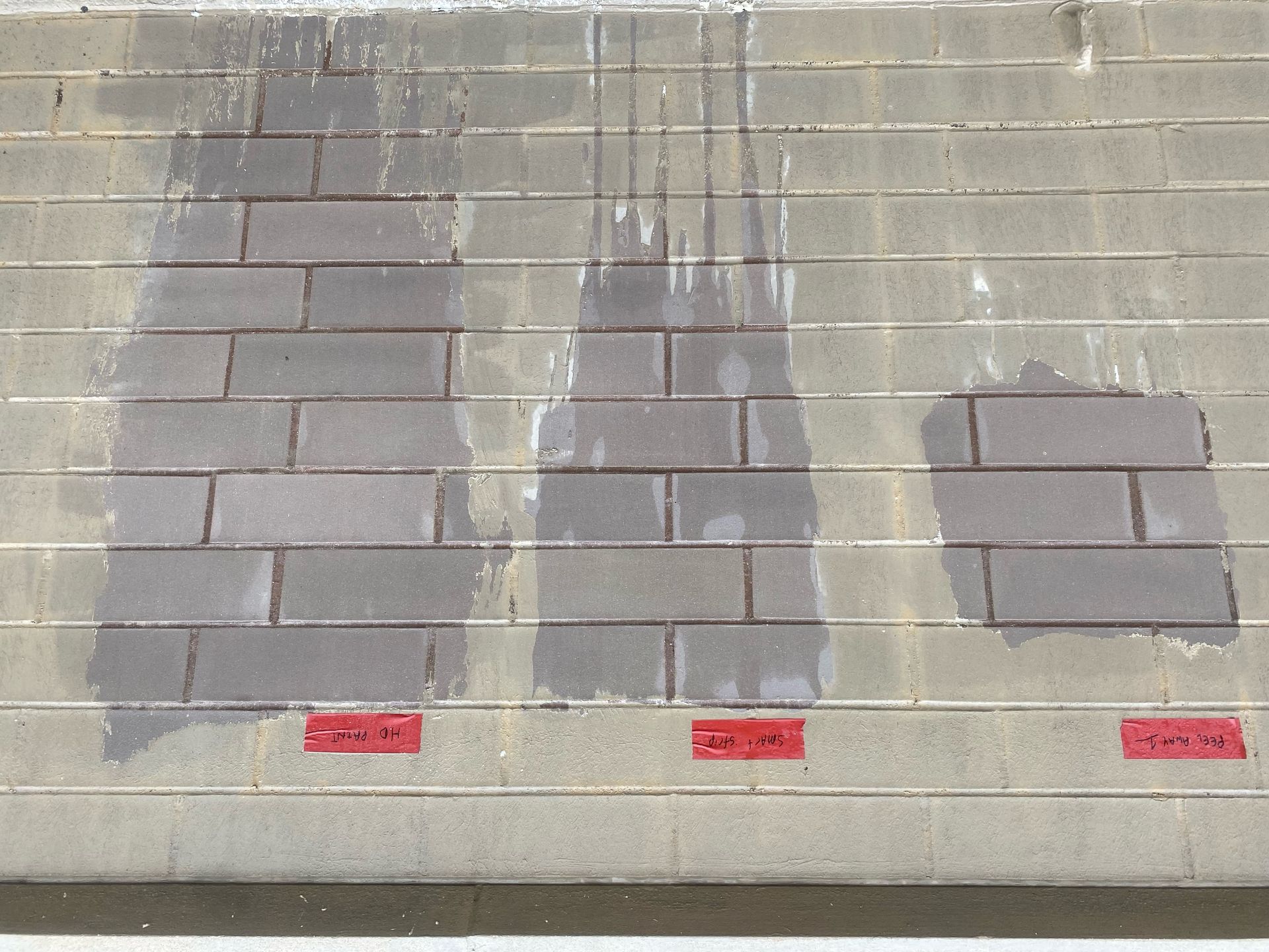 A masonry wall with multiple test panels evaluating different restoration methods for a cleaning project. The wall displays sections treated with various removal solutions, including Peel Away, Smart Strip, and HD Paint, with visible differences in effectiveness. Red tape labels beneath each panel identify the tested techniques. Some areas show partial paint or coating removal, while others exhibit streaking or residue, highlighting the trial process for selecting the best restoration approach.