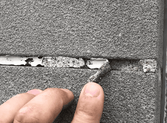 Close-up of a hand pulling away deteriorated sealant from a joint in a textured masonry wall. The old sealant is cracked and peeling, revealing gaps that could lead to water infiltration and structural issues. The image highlights the need for proper maintenance and replacement of failing sealants to protect the integrity of the building.