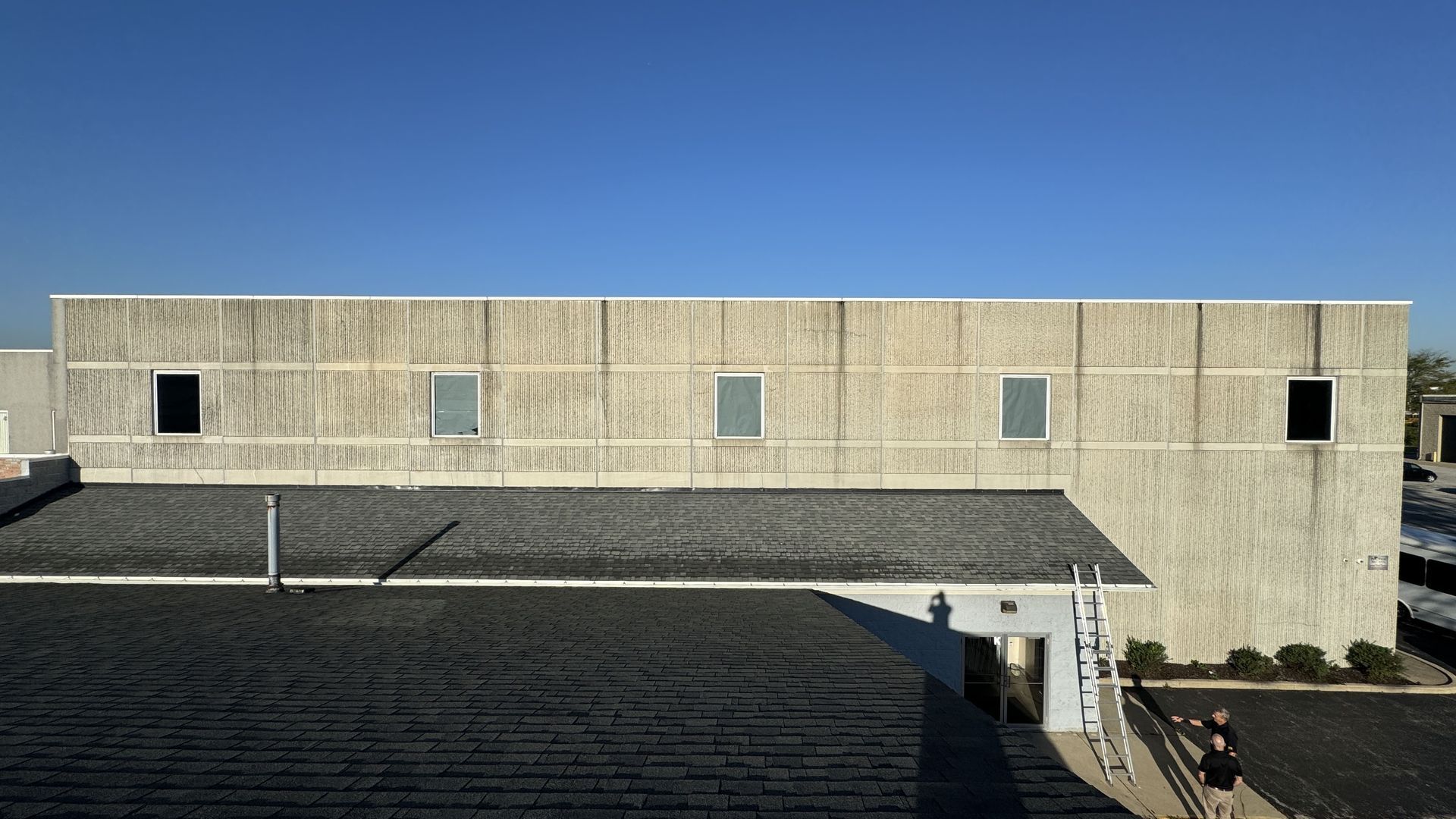 An exterior view of a building with a textured beige wall, five small windows, and a dark shingled roof under a blue sky.